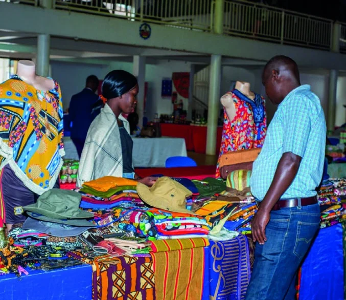 An exhibitor and visitor discuss colourful clothing and textiles at an exhibition stand.